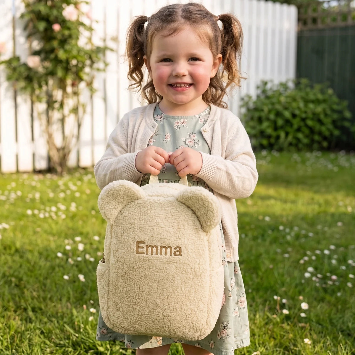 Mädchen hält den beigen Teddy Rucksack mit dem Namen „Emma“ im Garten